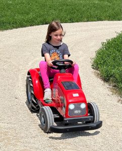 Young girl driving a red pedal tractor along a gravel path