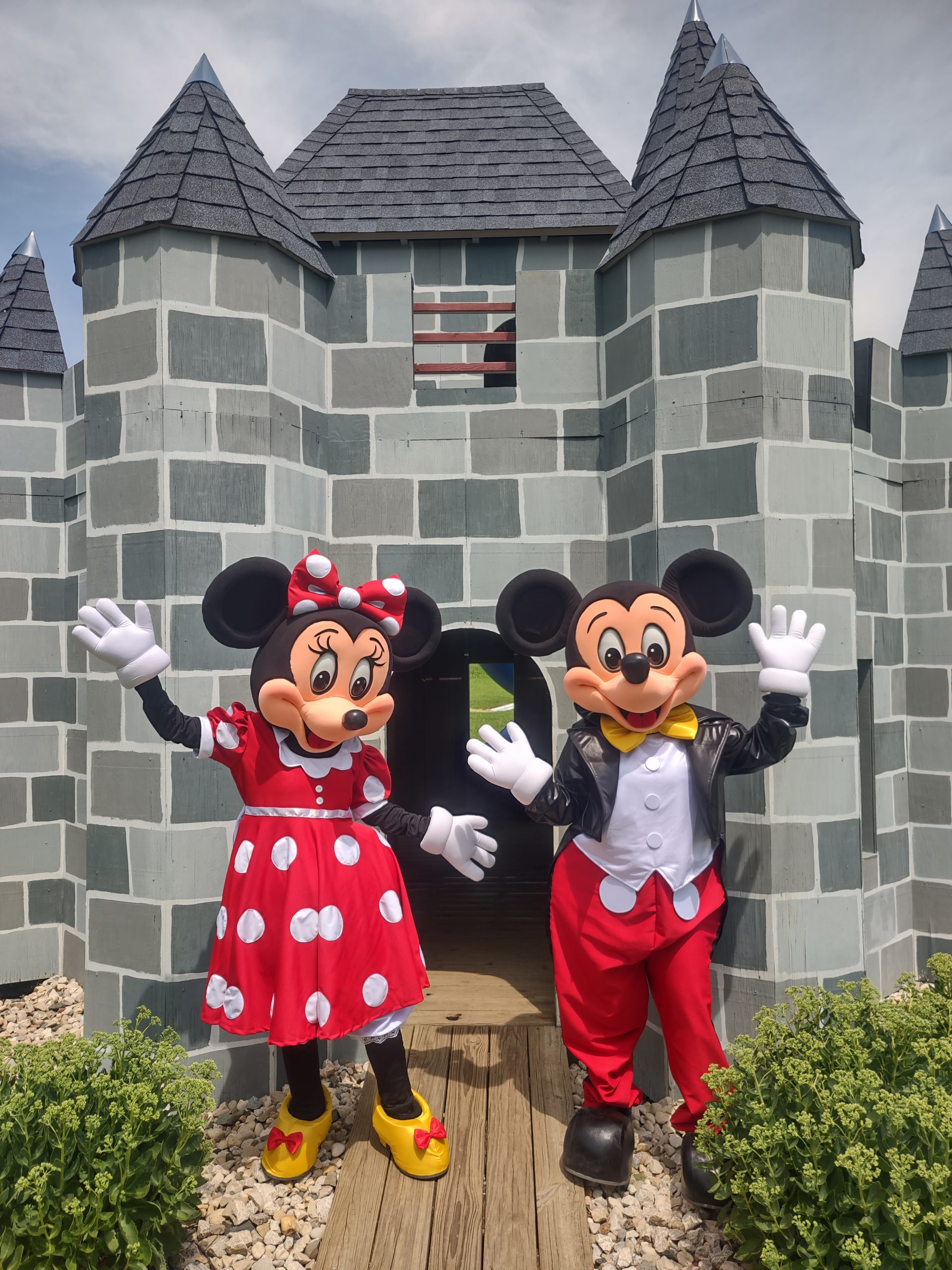 Mickey and Minnie Mouse characters waving in front of a gray play castle